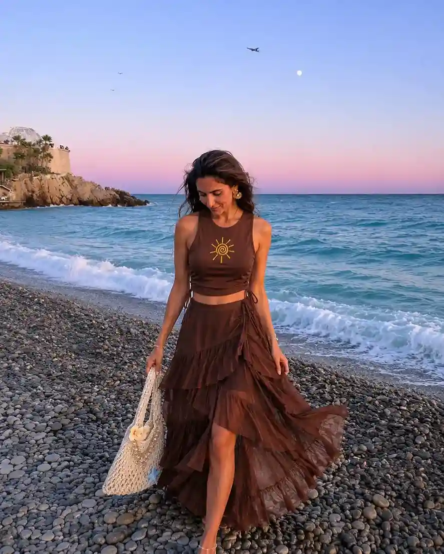Woman wearing a brown crop top and layered ruffle maxi skirt with a woven beach bag for a tropical beach vacation outfit.