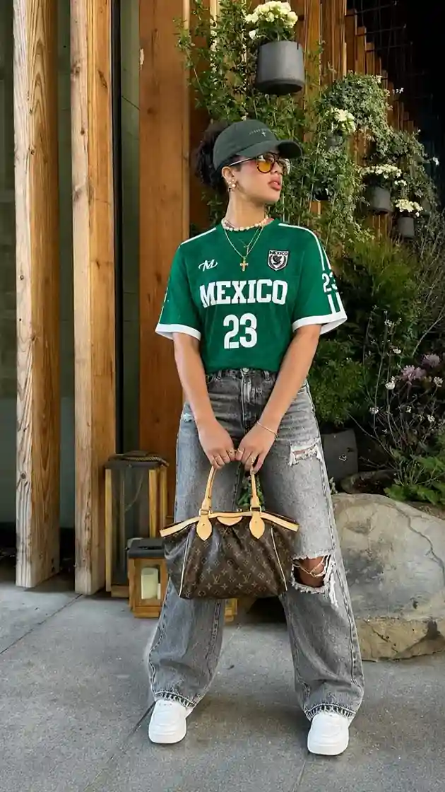 Woman wearing a casual mexico soccer game outfit with a green jersey, distressed straight-leg jeans, and a baseball cap.