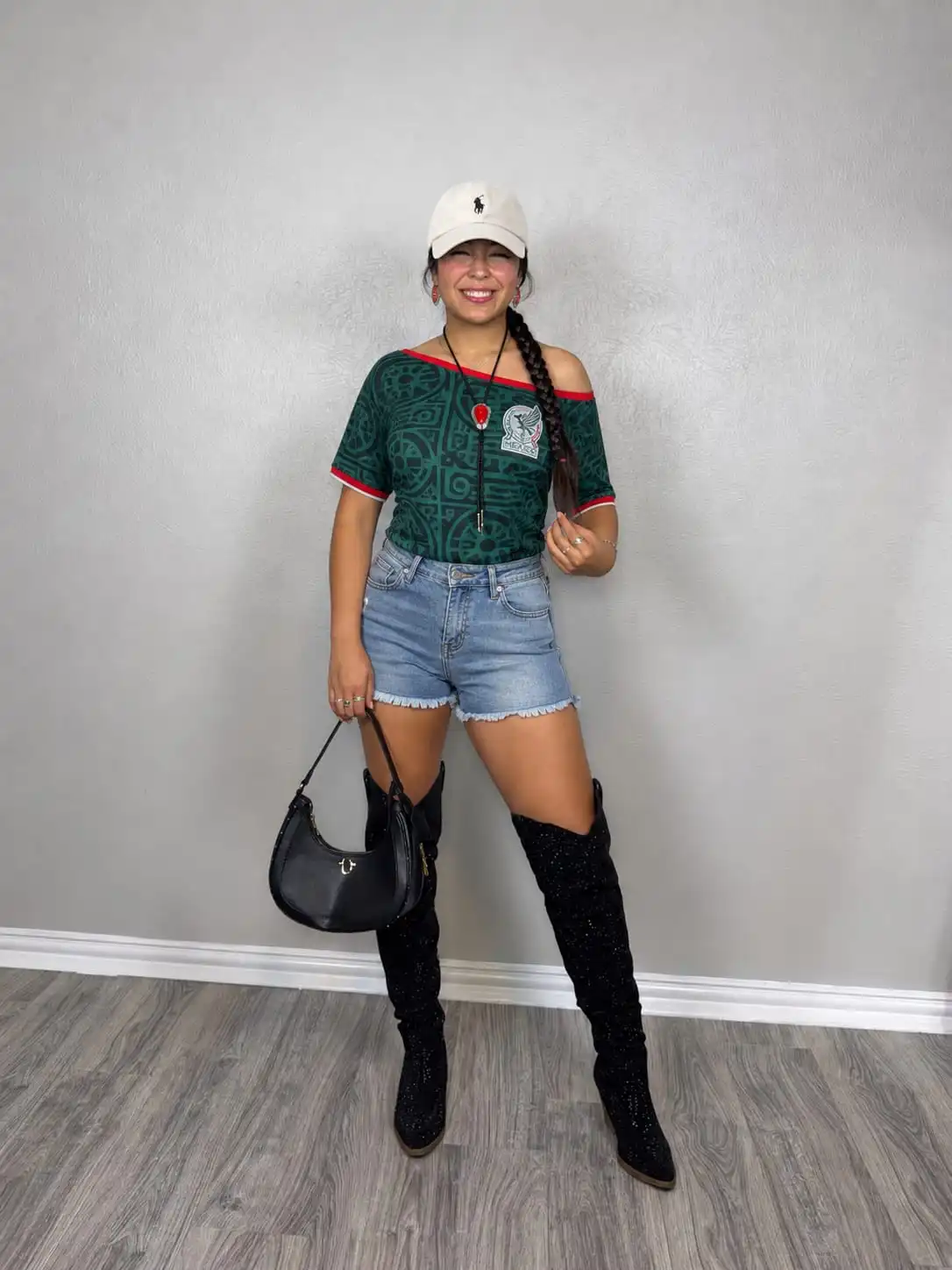Woman wearing a cute mexico game outfit with an off-shoulder Mexico jersey, light wash denim shorts, and a white baseball cap.
