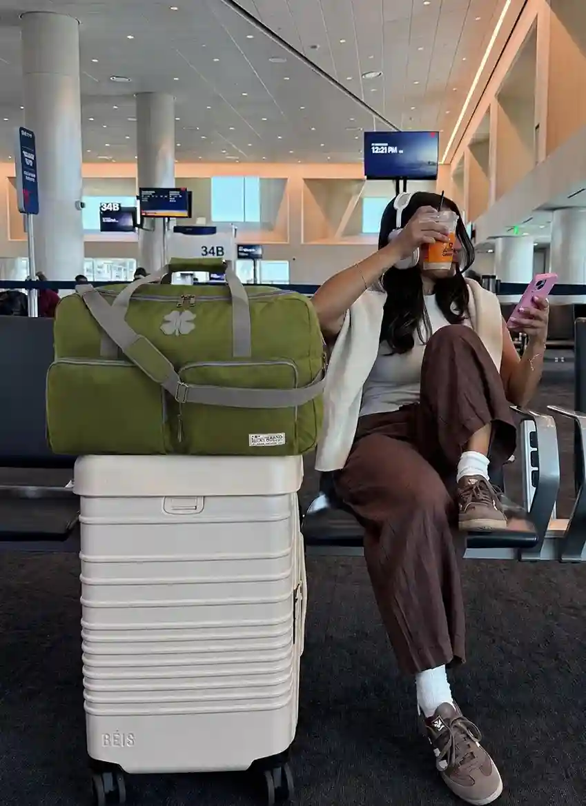 Woman waiting at an airport with a large green travel duffel bag placed on a suitcase, highlighting one of the best travel duffel bags for women with extra space for flights, weekend trips, and carry-on travel.