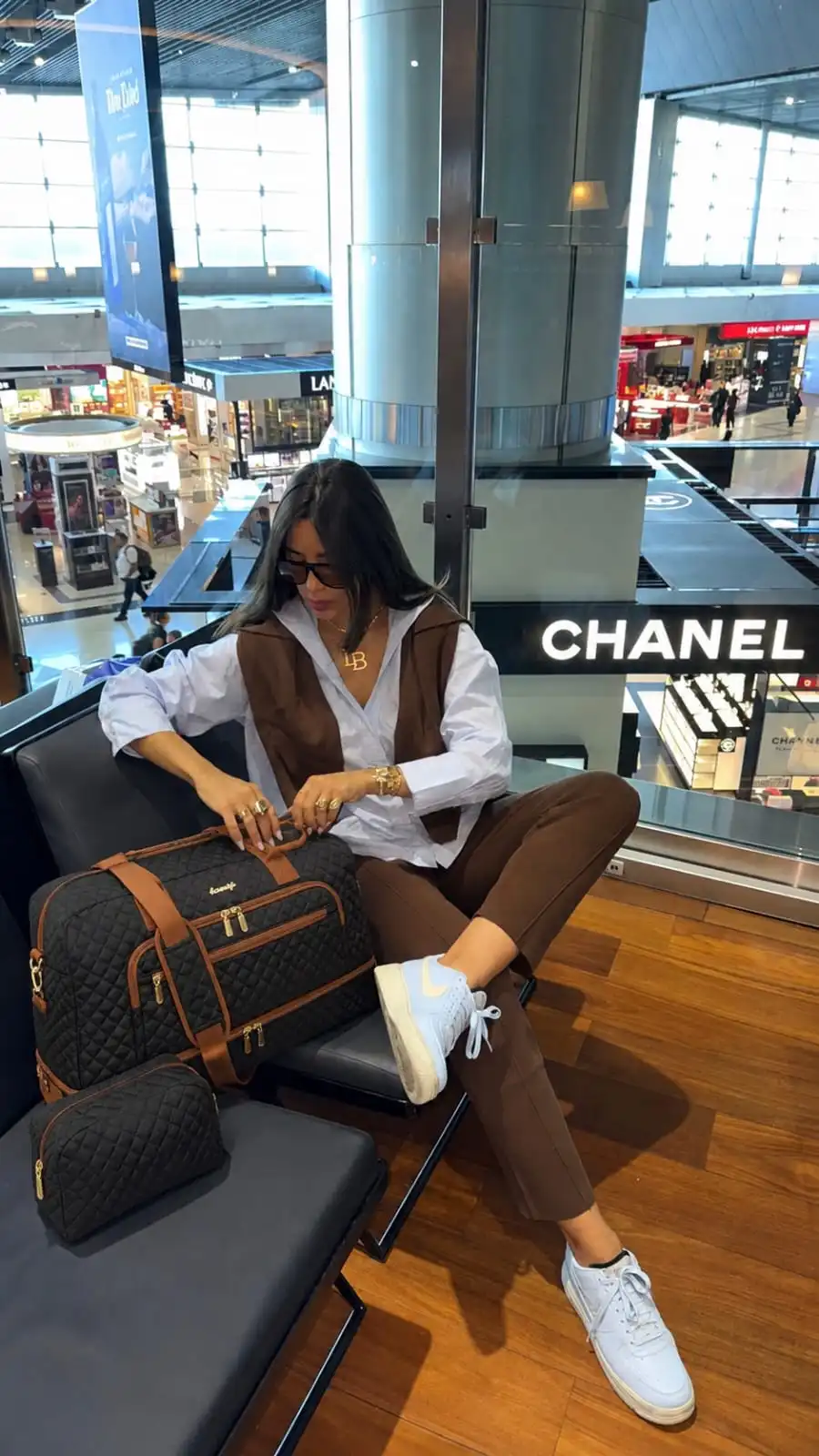 Woman sitting in an airport lounge with a black quilted travel duffel bag, featuring multiple compartments and extra storage space, ideal for weekend trips, carry-on travel, and organized packing.
