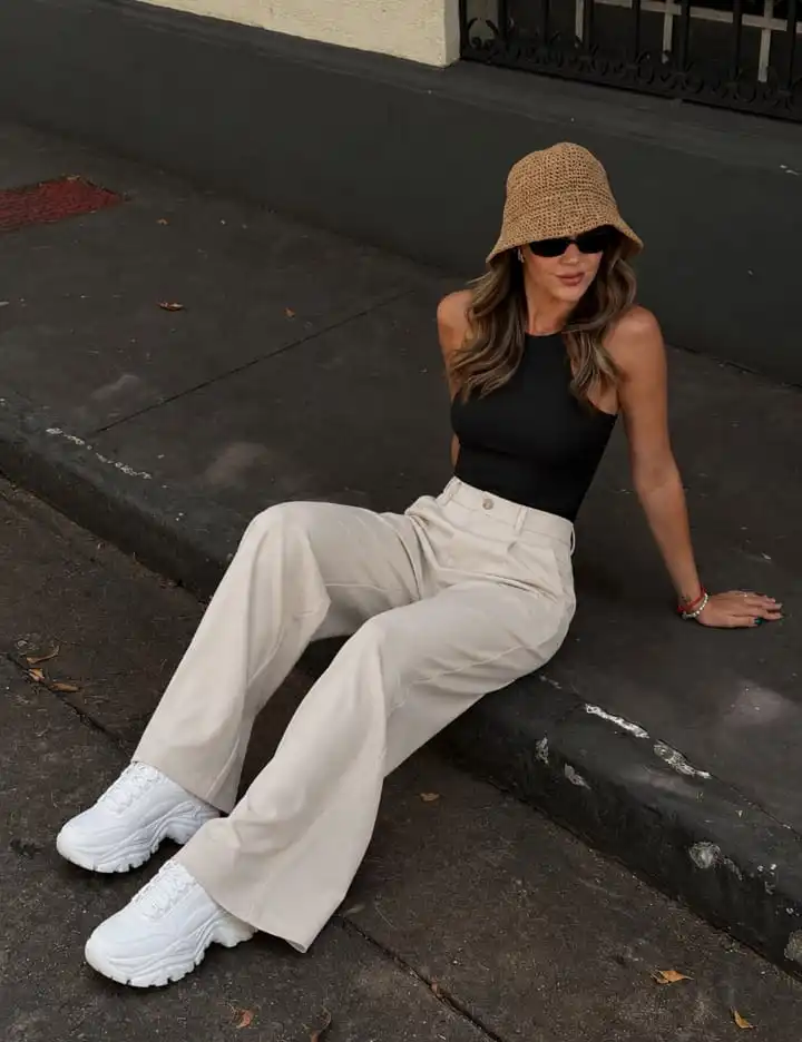 Woman wearing a black sleeveless bodysuit with beige wide-leg trousers and chunky white sneakers for a modern western outfit.