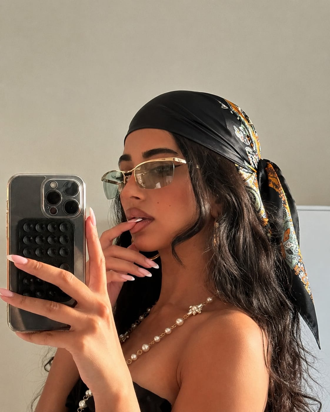 Woman wearing a black glossy satin paisley bandana with gold-rimmed glasses, pearl necklace, and a black strapless top, showcasing a stylish silk hair scarf for modern everyday fashion.
