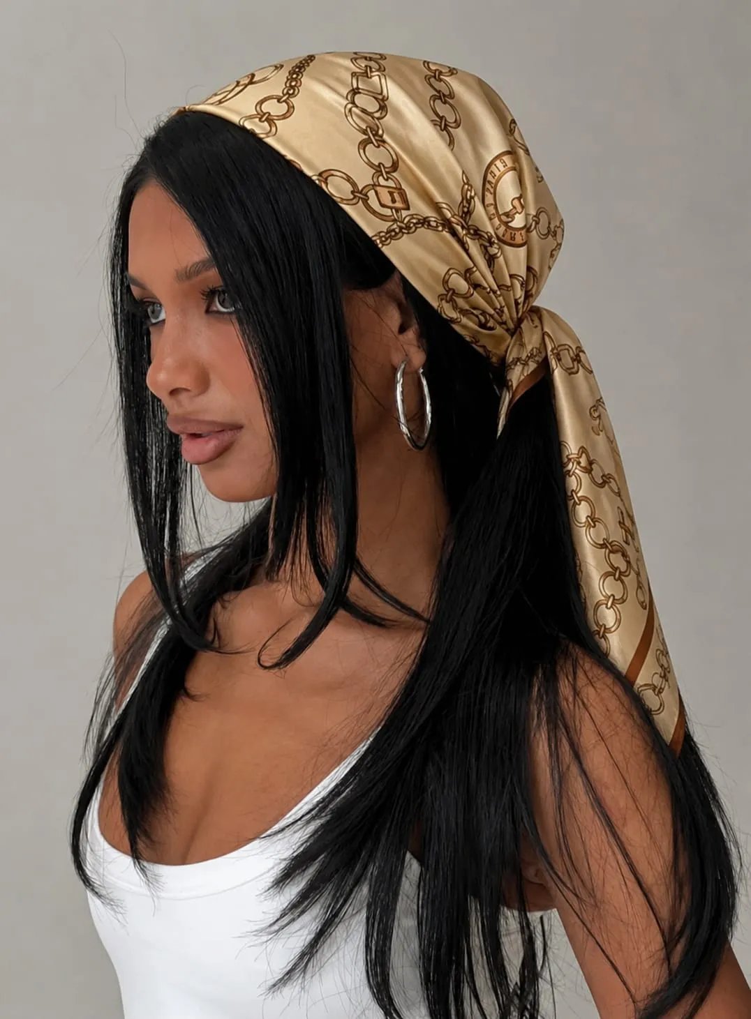 Woman wearing a beige and brown chain print glossy satin bandana styled as a silk head scarf with a white tank top, featuring an elegant Amazon hair scarf for casual, travel, and everyday outfits.