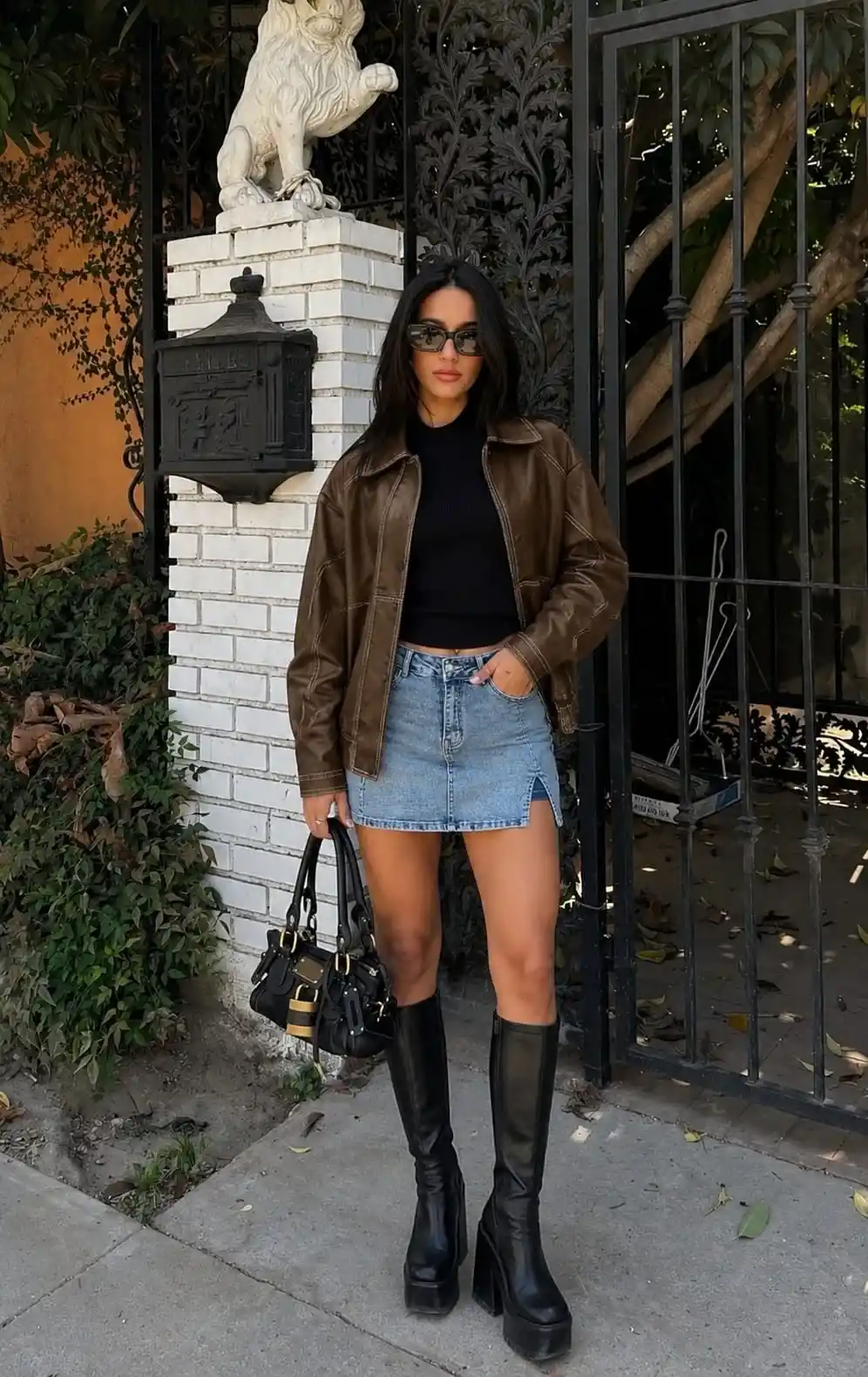 Woman wearing a brown leather jacket with a black knit top, denim mini skirt, and black knee-high boots for a stylish brown leather jacket outfit in 2026.