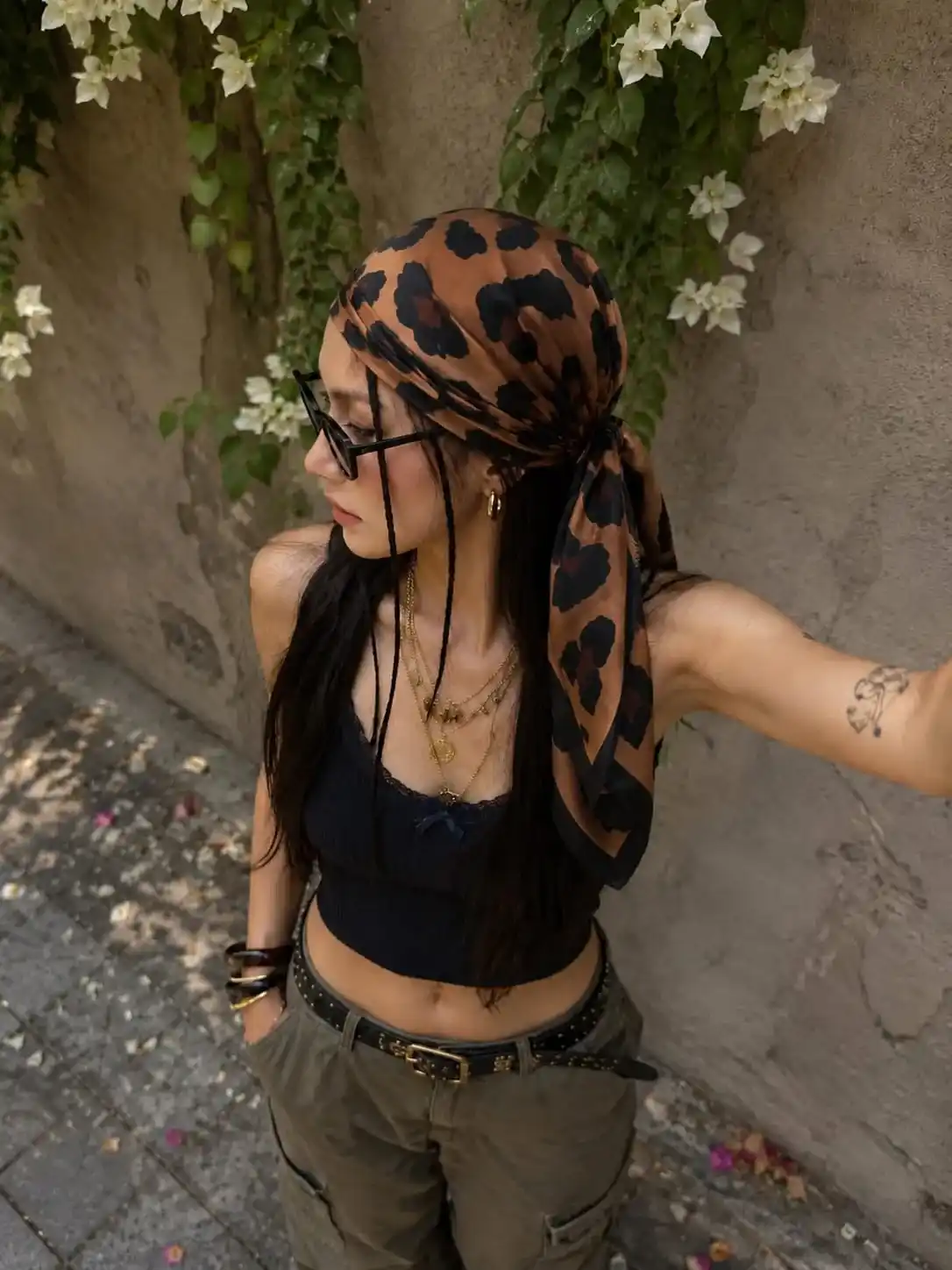 Woman wearing a glossy brown leopard print satin bandana with a black knit crop top, cargo pants, layered gold jewelry, and sunglasses, creating a fashionable street-style look with a silky hair scarf.