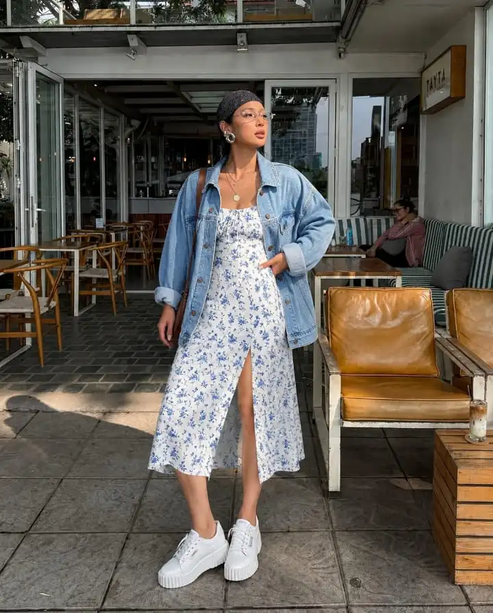 Woman wearing a blue floral midi dress with an oversized denim jacket and white platform sneakers for a trendy western outfit.