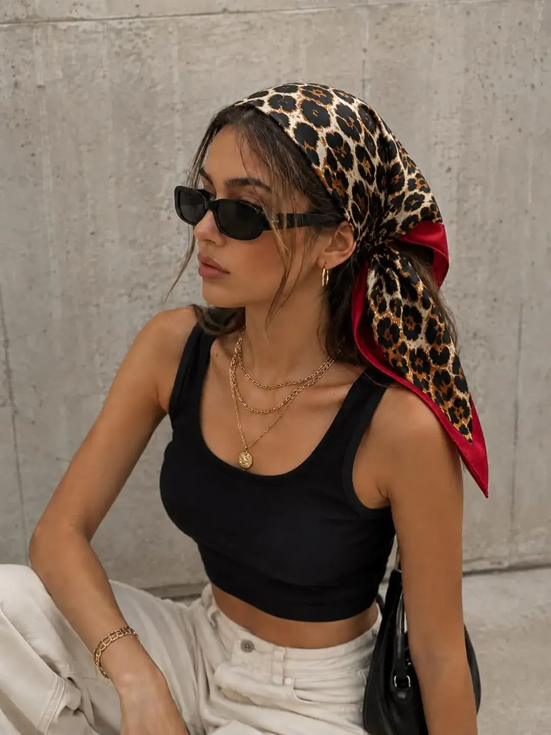 Woman wearing a glossy leopard print satin bandana with a black crop tank, white pants, layered gold necklaces, and sunglasses, showcasing a trendy silk hair scarf for casual everyday fashion.
