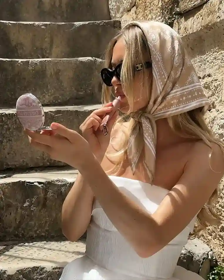 Woman wearing a neutral glossy satin bandana tied under the chin with a white strapless summer dress and black sunglasses, creating an elegant vacation-inspired look with a silky head scarf.