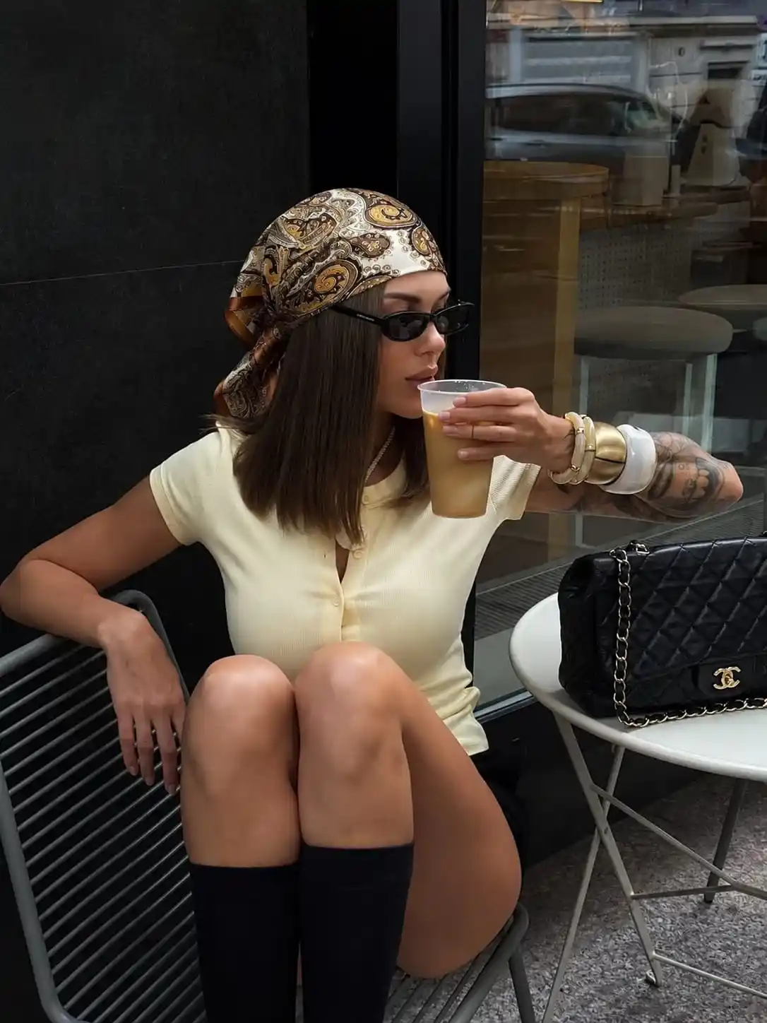 Woman wearing a glossy vintage paisley satin bandana with a cream knit top, black sunglasses, and gold accessories while enjoying coffee, showcasing a trendy silk head scarf for casual everyday style.